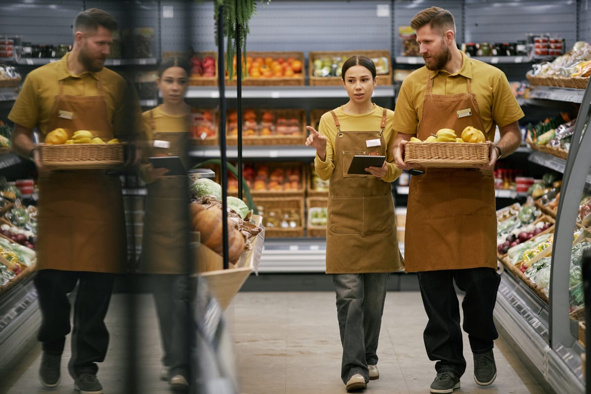 Two grocery associates in aprons walking a produce aisle together — one gesturing while holding a tablet, the other carrying a basket of fruit.