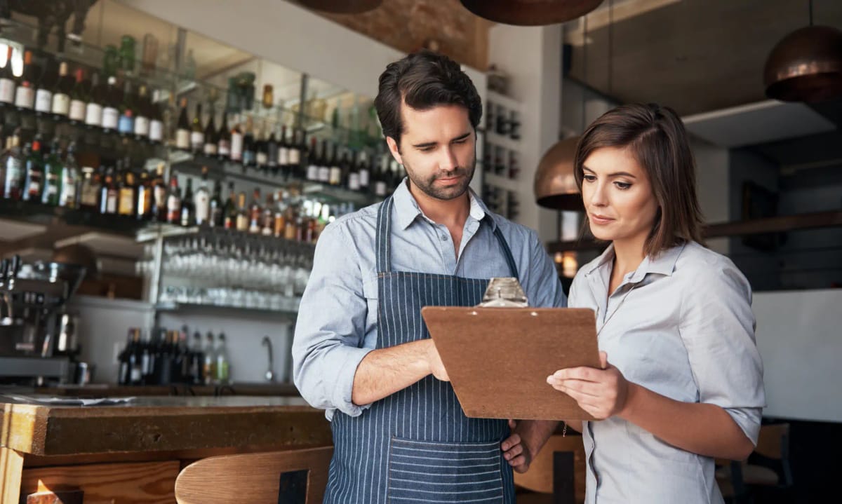 Two field leaders standing at a restaurant bar, reviewing notes together on a clipboard during a visit.