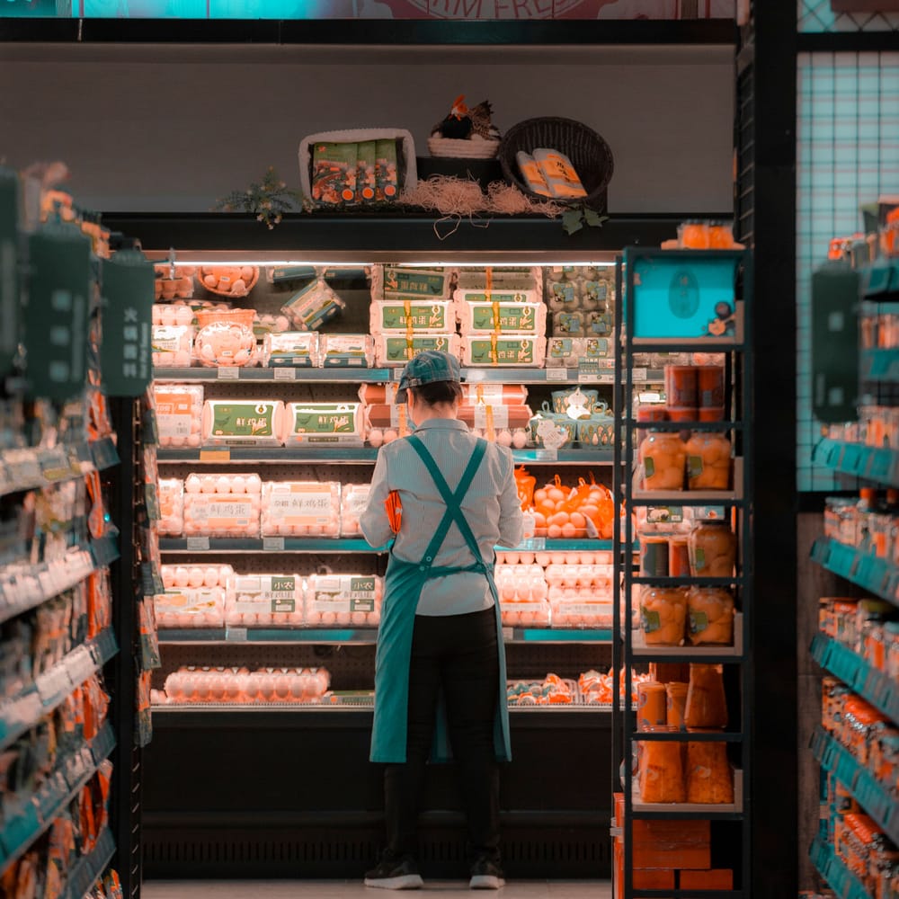 A grocery associate in an apron facing a refrigerated case of eggs and produce, restocking a store aisle.