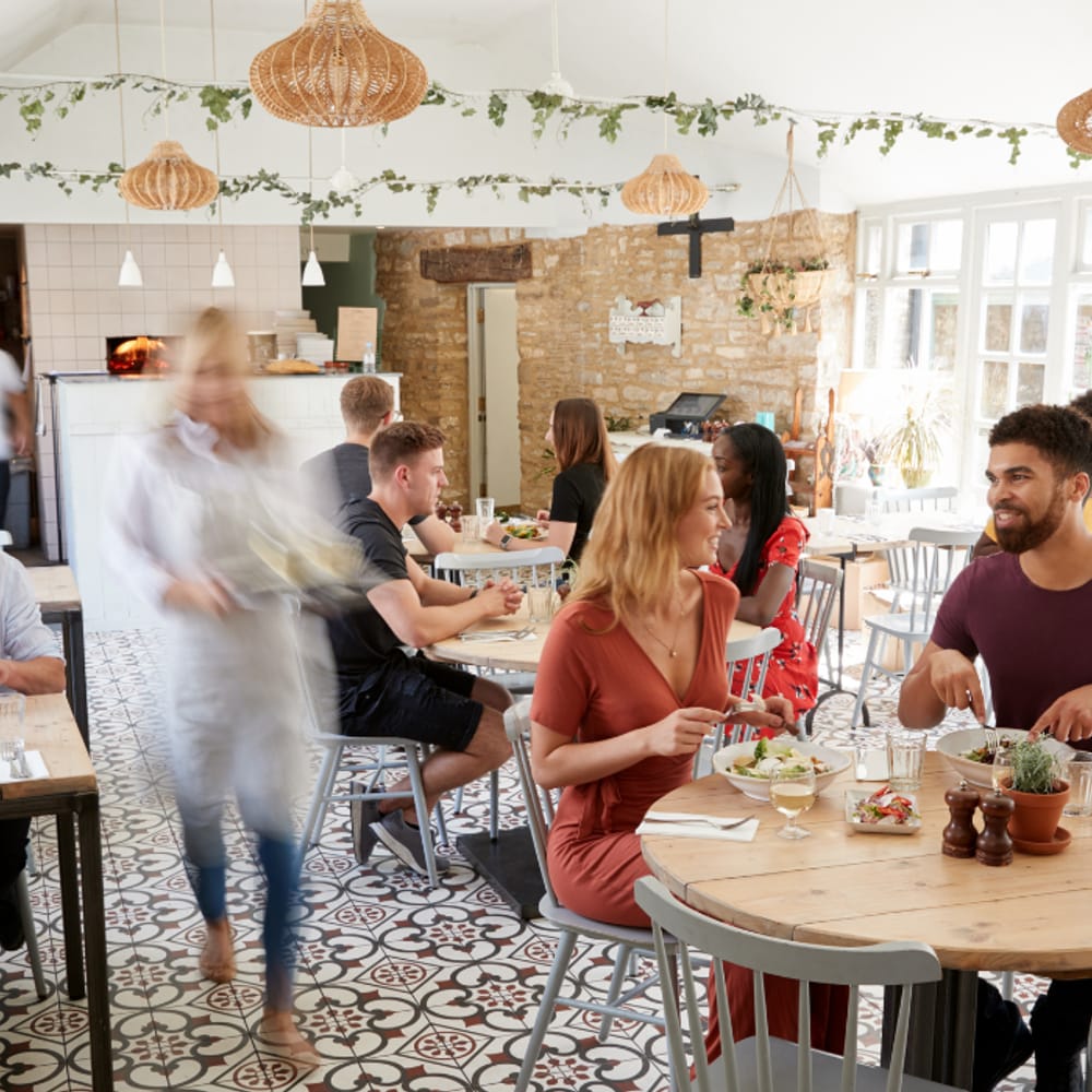 A busy sit-down restaurant floor at lunch — diners eating at wooden tables while a server crosses the room.