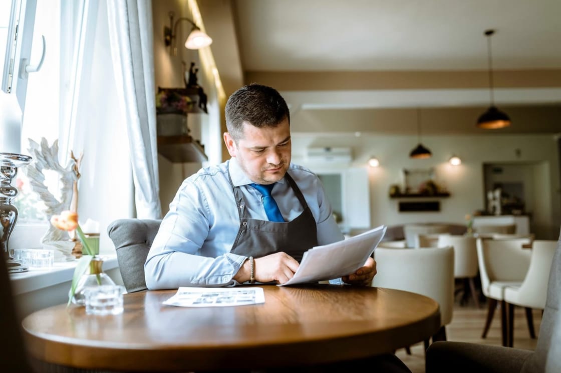 A restaurant manager sits alone at a table by a window, quietly working through printed reports and paperwork.